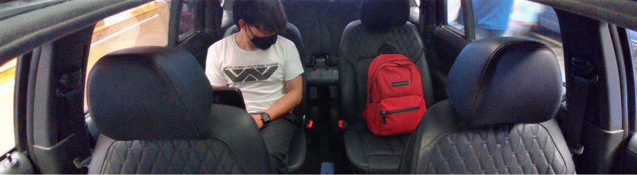 Outpainted D435-style frame from inside the BMW iX cabin: a passenger in a Weyland-Yutani t-shirt and black mask sitting in the rear seat with hands in lap; the cabin scene &mdash; quilted leather seats, headrests, door cards &mdash; extends out to both sides via outpainting.