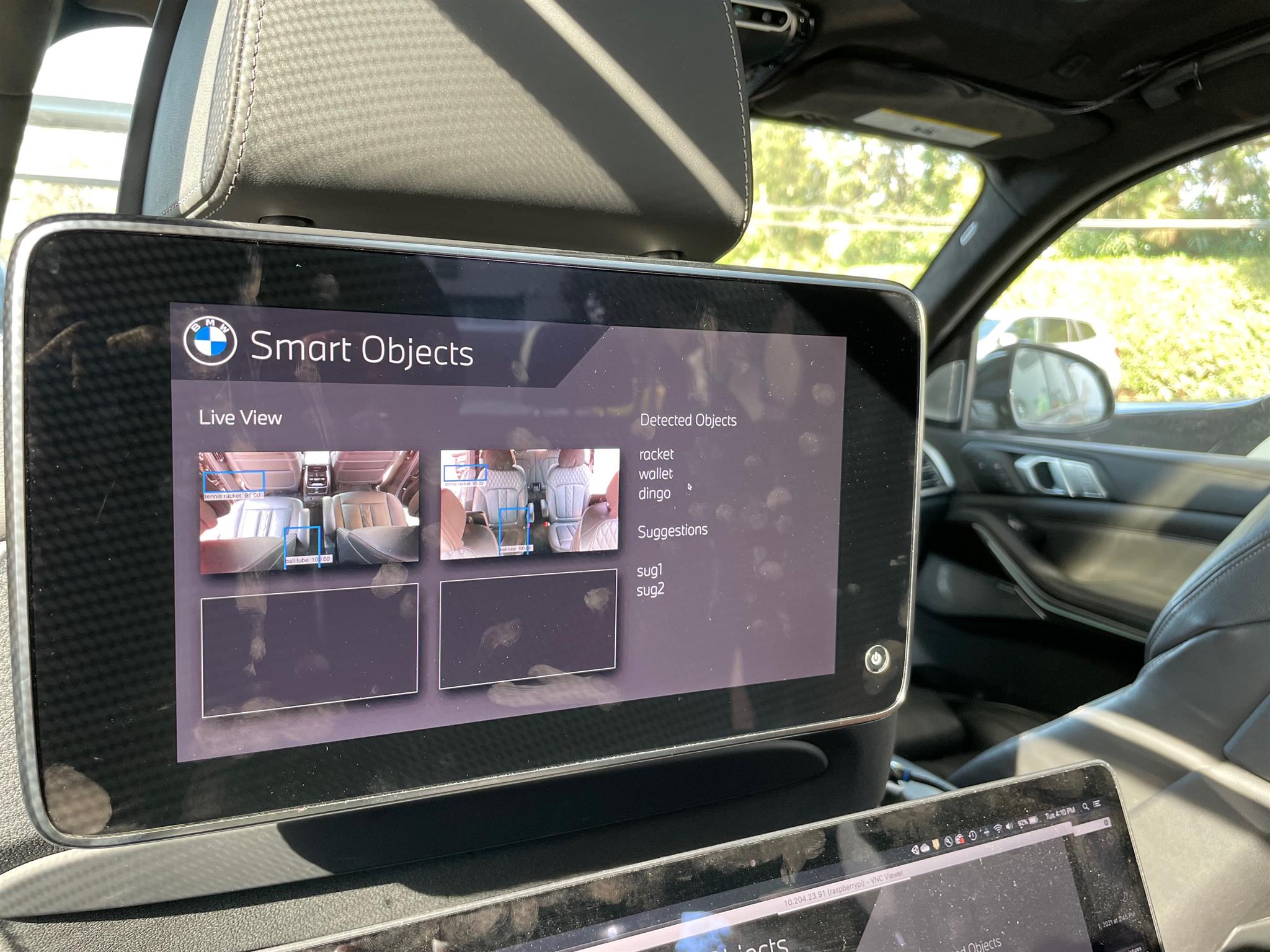 Running prototype on the BMW iX rear-seat head unit — Smart Objects screen showing Live View with bounding boxes around detected items in two camera tiles, a Detected Objects list (rocket, wallet, dingo), and a Suggestions panel below.