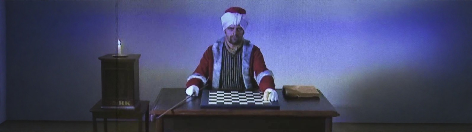 A grainy VHS-quality reenactment of the 18th-century Mechanical Turk &mdash; a man in a turban and fur-trimmed red coat seated at a chessboard against a blue-purple backdrop, in a dim wood-paneled study with a candle on a side table.